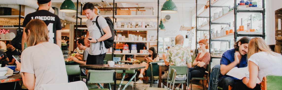 A busy cafe with people working and socializing. Some people are seated at tables with laptops, others are talking in groups. The cafe has a modern interior with green chairs and pendant lights hanging from the ceiling.